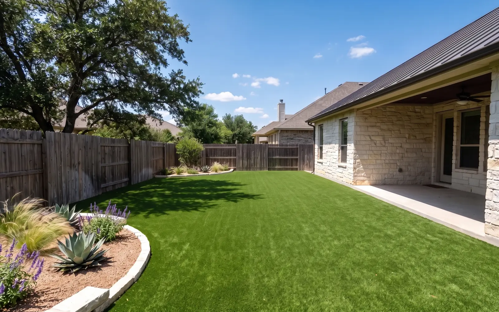 An artificial turf lawn behind a limestone house in Austin, with a live oak and a bed of agave and salvia